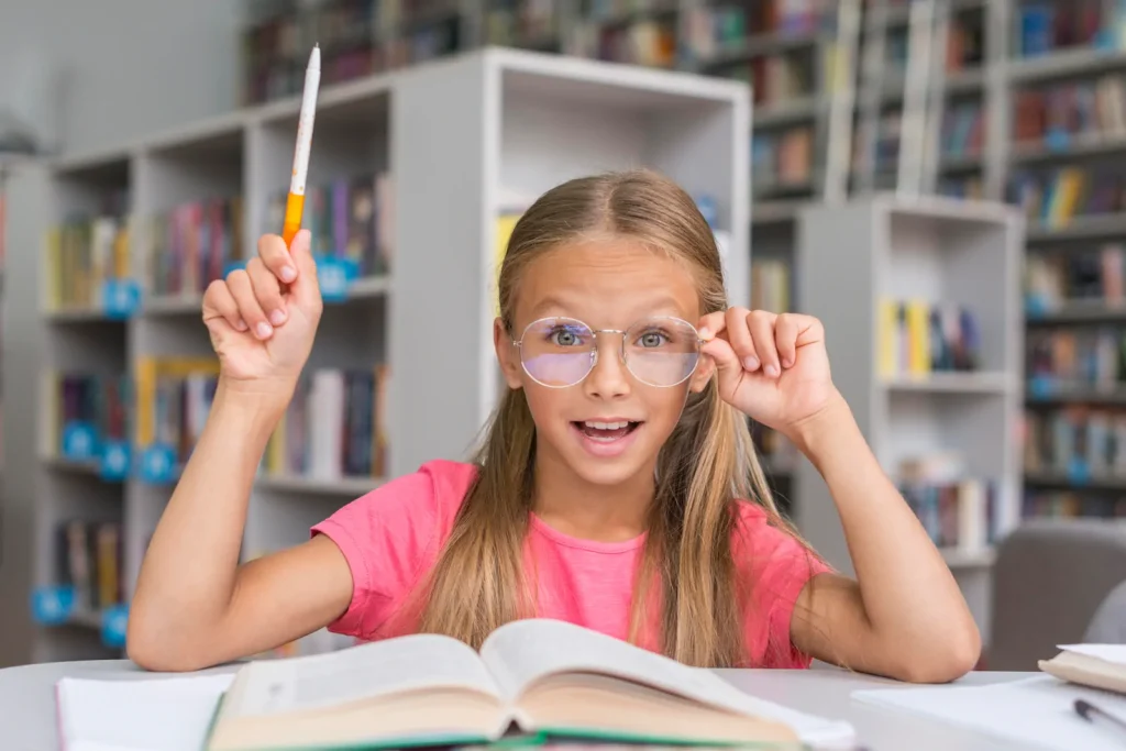 Niña con gafas levantando la mano mientras estudia un libro.