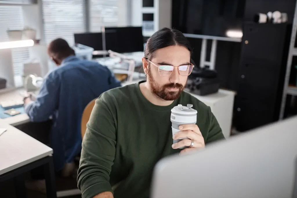 Profesional con barba usando computadora y sosteniendo una taza de café mientras usa lentes.