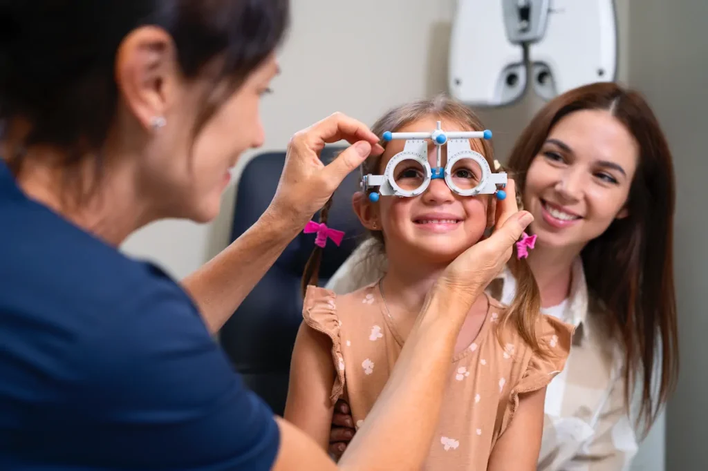 Niña realizando examen de visión con foróptero acompañada de su madre.
