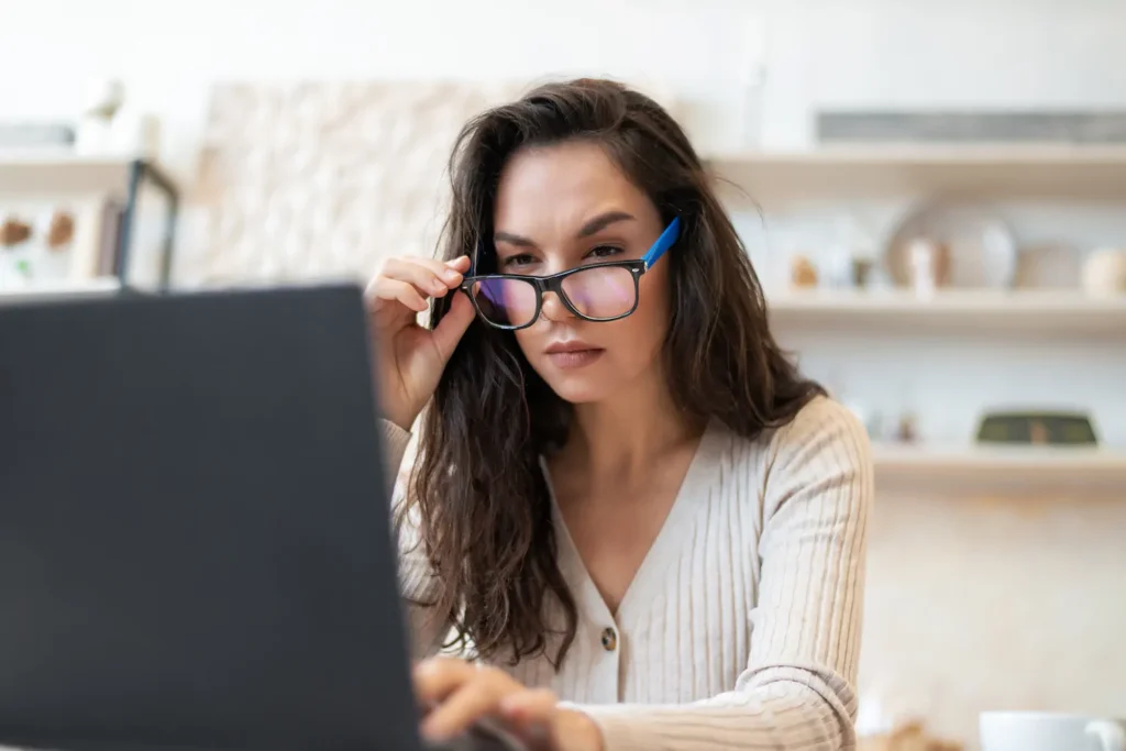 Mujer ajustando sus lentes frente a una laptop, posible indicio de fatiga visual por pantallas.