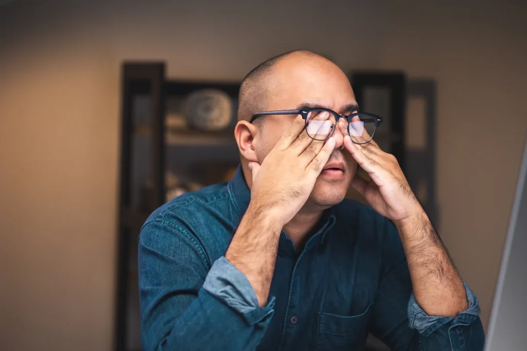 Hombre con lentes tocándose los ojos por molestias visuales causadas por uso prolongado de pantallas.