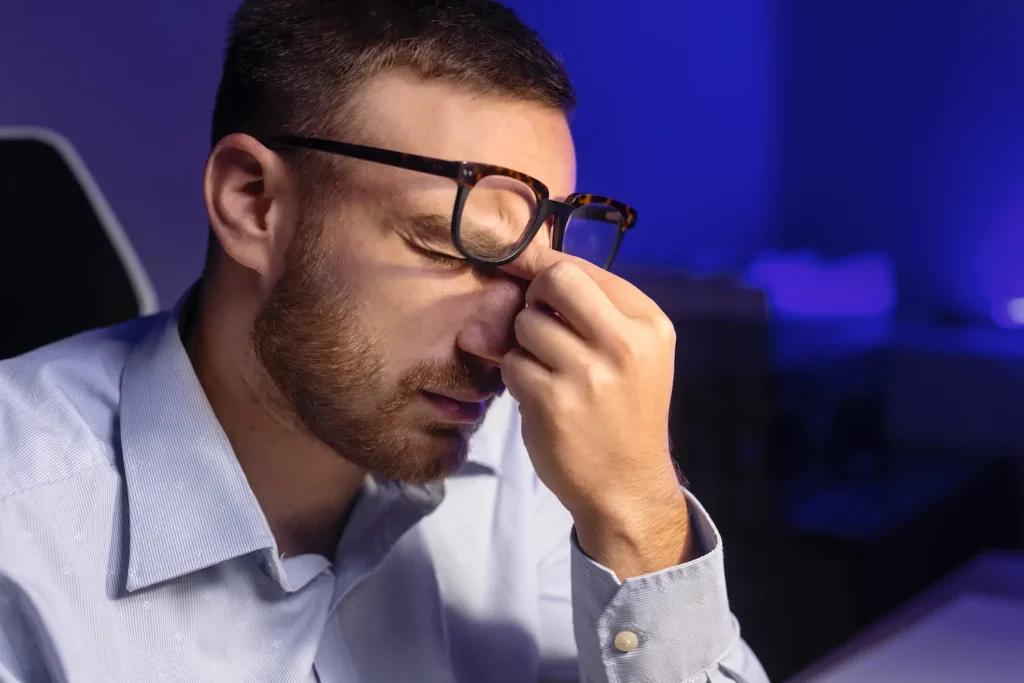 Hombre con lentes presionando el puente de la nariz mientras trabaja en computadora, síntoma de cansancio visual digital.