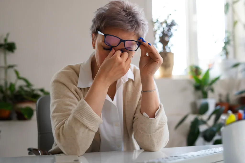 Mujer mayor frotándose los ojos por irritación y visión borrosa frente al computador.